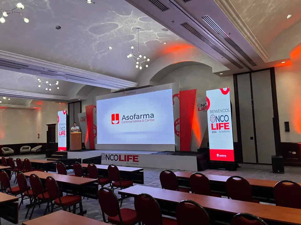 Escenario de OncoLife 2024 by Asofarma: tarima, marco blanco alrededor de la pantalla, piezas laterales rojas y tótems de bienvenida