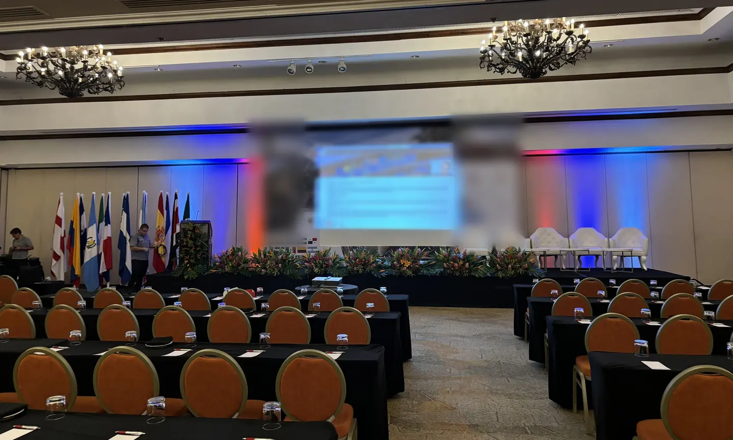 Salón plenario preparado para un simposio, con banderas, arreglos florales, escenario iluminado en azul y rojo y sillas en filas