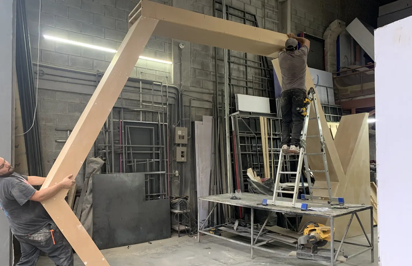 Dos operarios arman en el taller un pórtico de madera de gran formato: uno sostiene la pieza y el otro trabaja sobre una escalera